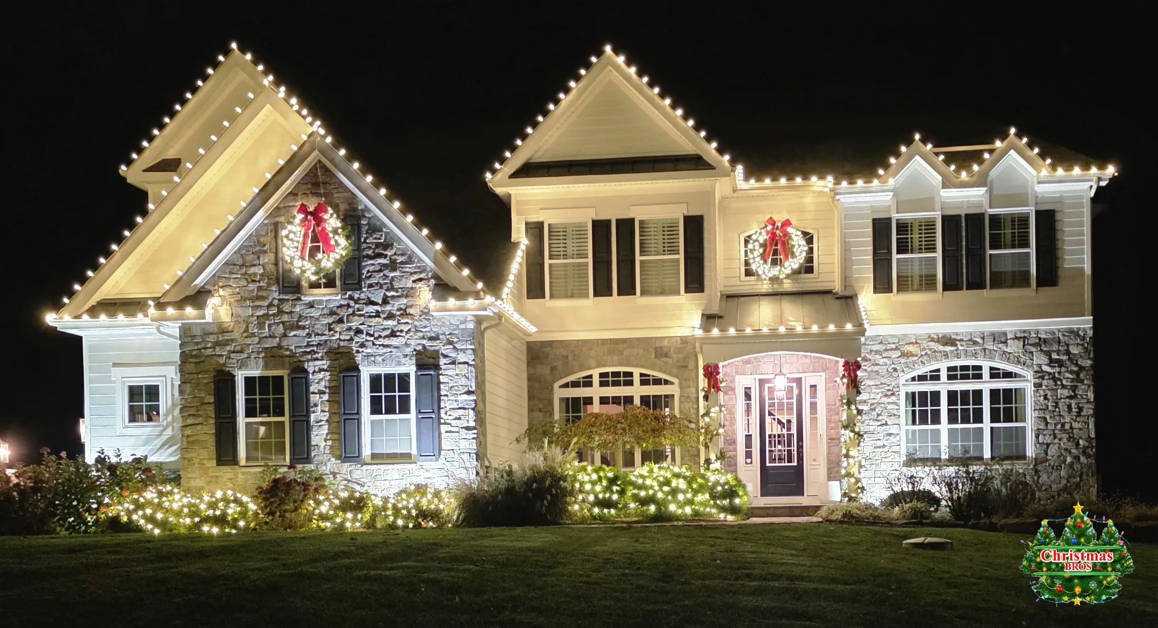 Stone two-story home in Chadds Ford, PA with warm white Christmas lights on every roofline and gable, wreaths and door garland, installed by Christmas Bros